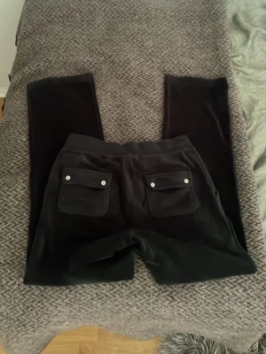 Juicy couture mjukisbyxor med fickor - Säljer ett par svarta mjukisbyxor i mjukt velourmaterial. Byxorna har stora fickor fram och bak, bakfickorna har lock med silverfärgade knappar. Resårmidja med dragsko för skön passform. Perfekta för chill dagar.