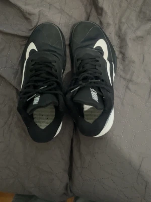 Svarta Nike sneakers med vit sula - Snygga svarta Nike sneakers med vit sula och vit swoosh på sidan. Skorna har lågt skaft och klassisk snörning. Ovandelen är i textil och syntetmaterial, och hälen har ett randigt mönster i svart och vitt. Perfekta för en sportig och avslappnad stil.