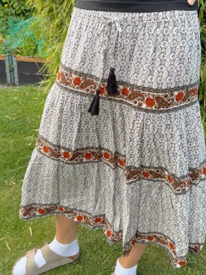 Mönstrad långkjol med volanger i boho-stil - Helt ny och oanvänd, superfin! Bohemisk långkjol med småblommigt mönster i vitt, svart och brunt. Kjolen har volanger, dekorativa band med orange och bruna detaljer samt snörning med tofsar i midjan. Perfekt för en avslappnad och trendig stil.
