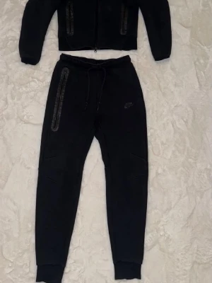 Nike tech tracksuit/dress S/M - Säljer ett par svarta mjukisbyxor från Nike med smal passform och ribbade muddar vid bensluten. Byxorna har dragkedjeförsedda fickor med detaljer i svart och justerbar midja med snörning. Perfekta för chill eller träning. Storlek small på tröjan medans medium på byxan nästan inget fel alls förutom ett litet skrap på de vänstra knäet men det syns knappt och färgen kan va lite of från när den va nyköpt den har bleknat pytte lite från solen