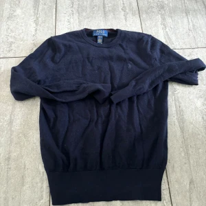 Mörkblå stickad tröja Polo Ralph Lauren L 14-16 år - Klassisk mörkblå stickad tröja från Polo Ralph Lauren med rund halsringning och diskret broderad logga på bröstet. Tröjan har långa ärmar och ribbade muddar vid ärmslut och nederkant. Perfekt för en stilren och tidlös look.