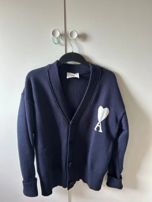 Mörkblå cardigan från Ami Paris - Mörkblå cardigan med knappar från Ami Paris. Tröjan har en vit hjärtlogga med bokstaven A på bröstet, ribbade muddar och en avslappnad passform. Perfekt för lager på lager och enkel att styla till olika outfits.