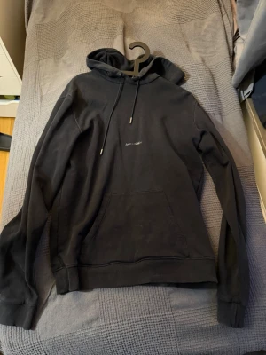 Saint Laurent hoodie - Säljer en svart hoodie från saint laurent med liten vit logga på bröstet. Klassisk modell med huva, dragsnören och stor känguruficka framtill. Ribbade muddar vid ärmslut och nederkant. Perfekt för en avslappnad streetwear-look.