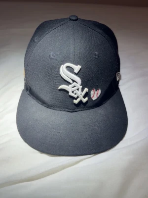 Svart Chicago White Sox keps New Era - Säljer en svart keps från New Era med Chicago White Sox-logga framtill och broderade detaljer som ett hjärta och texten 'LOVE & HUSTLE GAME' på sidan. Kepsen har platt skärm och broderad världsmästarlogga på sidan. Perfekt för dig som gillar streetwear.