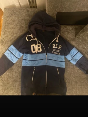 Blå DIF hoodie med dragkedja - Mörkblå hoodie med ljusblå ränder och vit text från DIF. Stor huva med vita snören, dragkedja framtill och två fickor med dragkedja. Tryck med '08' och 'D.I.F. EST. 1891 STOCKHOLM' på bröstet. Perfekt för dig som gillar sportig stil.