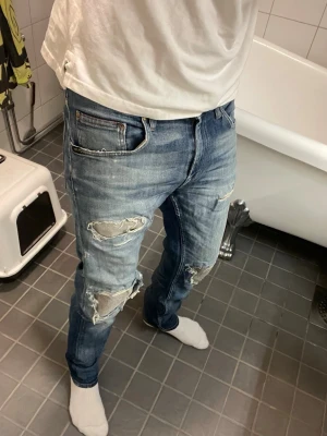 Blå slitna jeans från Tiger of Sweden - Säljer ett par blå jeans från Tiger of Sweden med slitningar och hål på knäna. Jeansen har en klassisk femficksmodell, normal midja och smal passform. Materialet är denim i bomull och de har en snygg tvättad look.