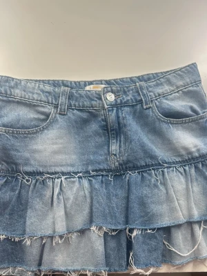 Blå jeanskjol med volanger - Säljer en kort jeanskjol i blå denim med två lager volanger och råa kanter för en trendig look. Kjolen har klassiska fickor fram och bak samt bälteshällor i midjan. Perfekt för dig som gillar en avslappnad men ändå cool stil.