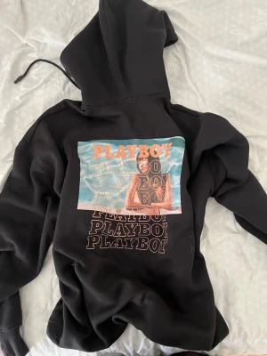 Svart hoodie från Playboy - Svart hoodie från Playboy med stor färgglad print på ryggen och flera PLAYBOY-tryck under. På bröstet finns en orange logga. Hoodien har huva med dragsko och är gjord i mjukt sweatshirtmaterial. Perfekt för dig som vill sticka ut med en streetwear-look.