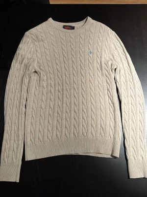 Beige kabelstickad tröja Morris - Snygg beige kabelstickad tröja från Morris i 100% pima cotton. Tröjan har rund hals, ribbade muddar och ett diskret blått broderat emblem på bröstet. Perfekt för dig som gillar klassisk och stilren design. 1800 nypris 