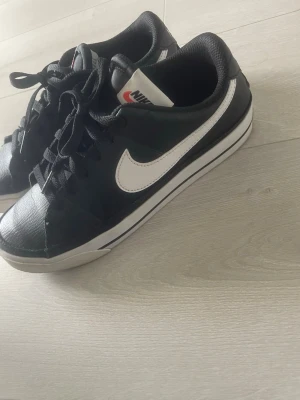 Svarta Nike sneakers med vit swoosh - Snygga svarta Nike sneakers med klassisk vit swoosh på sidan och vita detaljer på sulan. Skorna har lågt skaft, svarta skosnören och en bekväm, vadderad insida. Perfekta för dig som gillar en clean och sportig look.