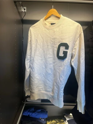 Beige sweatshirt från GANT - Vit sweatshirt i mjukt material med ribbade muddar vid ärmslut och nederkant. Stor grön bokstav 'G' i filt på bröstet ger en collegekänsla. Rund halsringning och avslappnad passform, perfekt för en casual look. Knappast använd endast vid ett fåtal tillfällen.