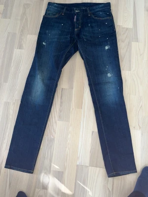 Mörkblå slim fit jeans Desquared  - Säljer ett par mörkblå slim fit jeans från Desquared med slitningar och färgstänk för en cool, använd look. Klassisk femficksmodell med kontrastsömmar och läderpatch bak i midjan. Perfekta för dig som gillar streetwear och en avslappnad stil.