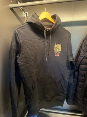 Svart hoodie från Morris - Snygg svart hoodie från Morris med guldfärgat tryck och brittisk flagga på bröstet. Klassisk känguruficka framtill och justerbar huva med snören. Tillverkad i mjukt bomullsmaterial som är skönt att ha på sig. Perfekt för en avslappnad och stilren look. Bra skick