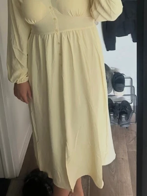 Ljusgul långklänning med knappar - Fin ljusgul långklänning med långa ärmar och knappar framtill. Klänningen har en markerad midja och lätt v-ringad halsringning. Perfekt för sommarens alla tillfällen.