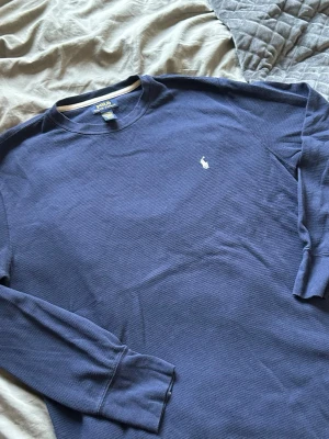 Blå långärmad tröja från Polo Ralph Lauren - Mörkblå långärmad tröja från Polo Ralph Lauren med klassisk vit broderad logga på bröstet. Tröjan har rund halsringning och är tillverkad i ett mjukt, strukturerat bomullstyg som ger en avslappnad men stilren look.