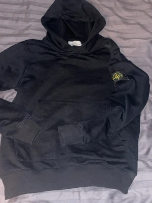 Svart hoodie från Stone Island - Säljer en svart hoodie från Stone Island med klassisk huva och känguruficka. Ikonisk patch med logotyp på vänster ärm. Mjuk bomullskänsla och ribbade muddar. Perfekt för dig som vill ha en stilren och trendig look.