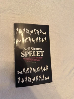 Spelet av Neil Strauss - En svart pocketbok med titeln 'Spelet' av Neil Strauss. Omslaget har vita siluetter av människor i olika poser och text i vitt och rött. Boken handlar om förförelse och relationer och är utgiven av Lind & Co. Oanvänd!