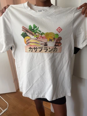 Casablanca T-shirt  - Vit t-shirt från CASA med färgstarkt grafiskt tryck av grönsaker och japansk text på bröstet. Klassisk rund hals och korta ärmar. Perfekt för dig som gillar unika prints och streetstyle.
