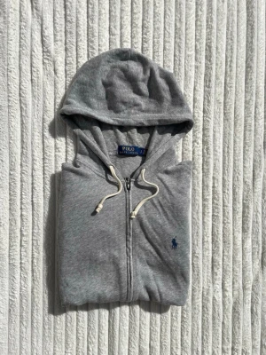 Grå zip hoodie från Polo Ralph Lauren - Snygg grå hoodie med dragkedja från Polo Ralph Lauren. Klassisk design med huva och vita dragsnören, två fickor framtill och den ikoniska blå loggan broderad på bröstet. Tillverkad i mjukt bomullsmaterial som är skönt att ha på sig.