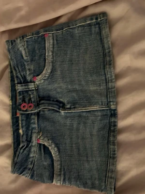 Kort jeanskjol💕💕 - Säljer en kort jeanskjol i blå denim med rosa knappar och en lila blomdetalj på ena bakfickan. Kjolen har klassiska fickor fram och bak samt markerade sömmar. Perfekt för dig som gillar en avslappnad och trendig stil. Har klippt av två fjärilar men syns knappt💕💕