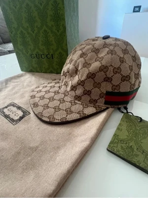 Gucci keps - Säljer en beige keps från Gucci i GG Supreme-mönster med klassiska gröna och röda ränder på sidan. Kepsen har böjd skärm och justerbar rem bak. Materialet är canvas med läderdetaljer. Perfekt för dig som vill ha en ikonisk accessoar med lyxig känsla, kepsen är helt ny och är alltid öppen för förslag!