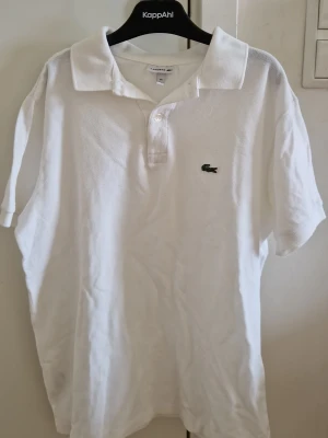 Vit piké från Lacoste med logga - Klassisk vit pikétröja från Lacoste med den ikoniska gröna krokodilloggan broderad på bröstet. Tröjan har korta ärmar, krage och knappar vid halsen. Tillverkad i mjuk bomull som känns skön mot huden. Perfekt för en clean och stilren look.