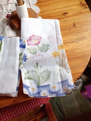 Köksgardinset med blommönster - Säljer ett köksgardinset i vitt tyg med färgglatt blommönster i rosa, gult, grönt och blått. Tyget har stora blommor och blad samt en blå bård med vita detaljer. Perfekt för att ge köket en retro och somrig känsla.