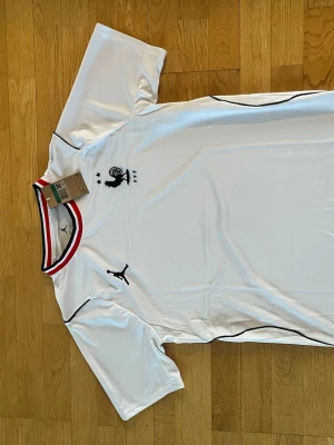 Vit Puma fotbollströja Frankrike - Säljer en vit fotbollströja från Puma med Frankrikes landslagsemblem och svarta detaljer. Tröjan har korta ärmar och en ribbad krage med röd, vit och blå rand. Perfekt för fotbollsträning eller match.