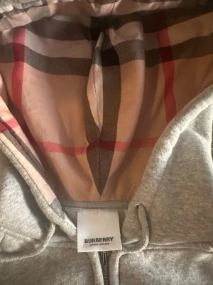 Grå zip-hoodie från Burberry - Snygg grå zip-hoodie från Burberry med klassiskt rutigt foder i huvan. Tröjan har dragsko i huvan, fickor framtill och dragkedja hela vägen. Perfekt för dig som gillar stilrena detaljer och ikoniska mönster. Funkar som size S och M