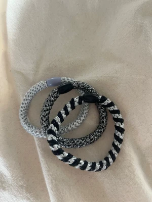 Set med tre elastiska hårsnoddar - Säljer ett set med tre elastiska hårsnoddar i svart, vitt och silver. Två av snoddarna har svart plastdetalj och en har ljuslila plastdetalj. Perfekta för att fixa håret eller använda som accessoar på handleden. Materialet är stretchigt tyg med flätad design.