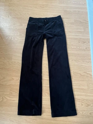 Svarta juicy byxor  - Säljer ett par svarta mjukisbyxor i mjuk velour med bred resår i midjan och snörning. Byxorna har raka ben och två fickor fram samt två bakfickor med lock och silverfärgade knappar. Perfekta för chill dagar eller när du vill vara extra bekväm.