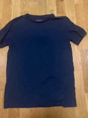 Blå t shirt - En enkel mörkblå t-shirt med rund hals och korta ärmar. T-shirten har en klassisk passform och är tillverkad i mjukt tyg, perfekt för en avslappnad stil. Inga synliga tryck eller mönster.