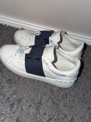 Vita Valentino sneakers med blå detalj - Säljer ett par vita sneakers från Valentino med bred mörkblå rem över snörningen och coola nitar på hälen. Skorna är i läder och har en clean, stilren look som passar till det mesta. Klassisk låg modell med vita snören och diskret logga på plösen. Skick 9/10! Nästan helt nya. Använda fåtal gånger. Hör av dig vid frågor eller funderingar! Inget og ingår tyvärr😊