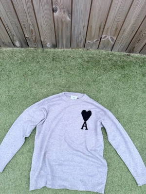 Grå stickad tröja från Ami Paris - Säljer en ljusgrå stickad tröja från Ami Paris med svart hjärta och bokstaven A broderat på bröstet. Tröjan har rund halsringning, långa ärmar och ribbade muddar vid ärmslut och nederkant. Perfekt för dig som gillar stilrena detaljer.