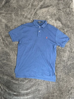 Ralph Lauren Blå piké - Klassisk blå pikétröja från Ralph Lauren med korta ärmar och krage. Tröjan har knäppning med två knappar och den ikoniska röda logotypen broderad på bröstet. Perfekt till jeans eller shorts för en clean look. Storlek M!