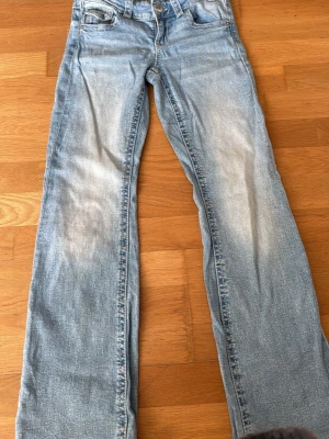 Ljusblå bootcut jeans med slitningar - Säljer ett par ljusblå bootcut jeans med snygga slitningar på framsidan och klassiska fickor bak med knappdetaljer. Jeansen har normal midja och är tillverkade i denim med en skön stretch. Perfekta för dig som gillar en avslappnad och trendig look.