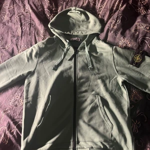 Grön stone island hoodie - Snygg grön hoodie från Stone Island med dragkedja framtill och klassisk logopatch på vänster ärm. Hoodien har justerbar huva med snören och två fickor framtill. Obs aldrig använt 
