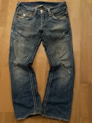 True religon jeans  - Säljer ett par blå jeans med raka ben och slitningar på både fram- och baksida. Jeansen har tydliga kontrastsömmar och bakfickor med dekorativa sömmar. Perfekta för en avslappnad och trendig look.
