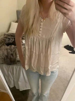 Beige blus med knappar och spetsdetaljer - Söt beige blus med rund halsringning och dekorativa knappar framtill. Blusen har korta ärmar med spetskant och ett luftigt, löst fall. Perfekt till jeans eller kjol. OBS! Jag kan stryka den innan jag skickar iväg den om de önskas🤍🤍