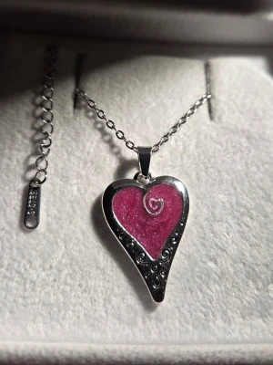 Halsband med hjärtformad lila berlock - Unikt handgjort halsband med hjärtformad berlock i lila och silver som lyser i mörker efter att ha varit i dagsljus eller solljus. Berlocken har en spiral i mitten och dekorativa detaljer längs kanten. Kedjan är i silverfärg och berlocken lyser i mörker med en cool blå-lila effekt. Perfekt för dig som gillar att sticka ut. Både hänget och kedjan tål vatten & missfärgas inte.
