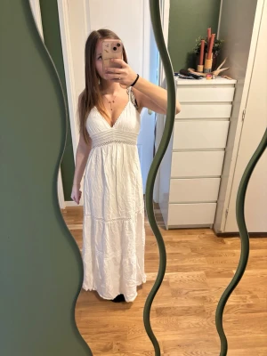 Vit långklänning med spetsdetaljer - Säljer en vit långklänning med smala axelband och v-ringning. Klänningen har fina spetsdetaljer och en lätt, luftig känsla som passar perfekt till sommaren. Materialet känns som bomull och modellen har en avslappnad passform.