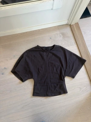 Brun topp med vida ärmar Gina Tricot - Säljer en brun topp från Gina Tricot i bomull med breda, korta ärmar och markerad midja. Toppen har en enkel, stilren design med söm framtill och rund halsringning. Perfekt för en chill och trendig look.