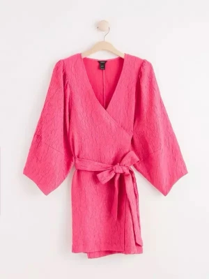 Rosa omlottklänning med knytband - Säljer en kortare rosa omlottklänning med v-ringning och vida ärmar. Klänningen har ett matchande knytband i midjan och en lätt strukturerad yta som ger extra känsla. Perfekt för dig som vill sticka ut med färg och stil.