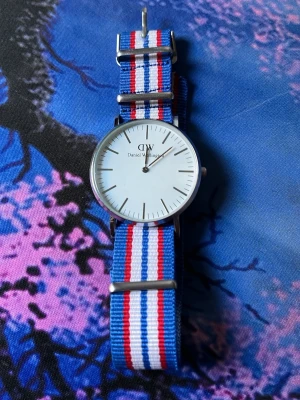 Daniel Wellington klocka med randigt band - Stilren klocka från Daniel Wellington med vitt ur och minimalistiska svarta markeringar. Klockan har ett tygarmband i blått, vitt och rött med silverfärgade detaljer. Perfekt accessoar för en clean och klassisk look.