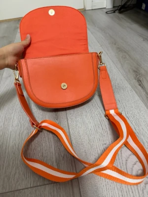 Orange axelväska med randigt band - Snygg orange axelväska i rundad form med magnetlås och justerbart axelband i orange och vitt. Väskan har guldfärgade metalldetaljer och är tillverkad i slätt konstläder. Perfekt för dig som vill ha en färgklick till outfiten.