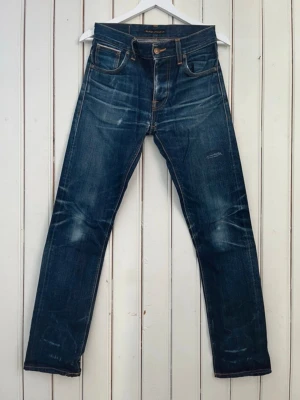 Blå jeans från Nudie Jeans - Säljer ett par klassiska blå jeans från Nudie Jeans med snygg slitning och kontrastsömmar. Jeansen har fem fickor, raka ben och en cool vintage look. Tillverkade i denim med detaljerade sömmar på bakfickorna. Perfekta för en avslappnad stil.