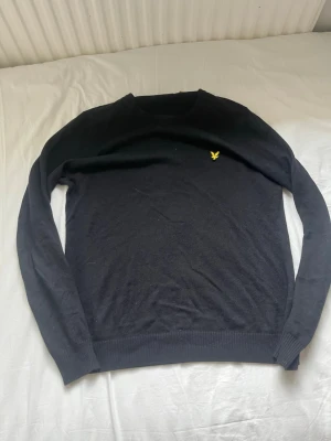 Svart stickad tröja från Lyle & Scott - Klassisk svart stickad tröja från Lyle & Scott med rund halsringning och den ikoniska gula örnloggan broderad på bröstet. Tröjan har långa ärmar och ribbade muddar vid ärmslut och nederkant. Perfekt för en stilren och enkel look.