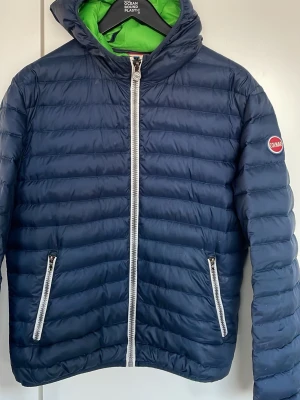 Mörkblå dunjacka från Colmar - Snygg mörkblå dunjacka från Colmar med vit dragkedja och två fickor framtill. Jackan har en huva med grön insida och Colmar-logga på ärmen. Perfekt för kalla vinterdagar och riktigt lätt att bära. Klassisk quiltad design och sportig look.