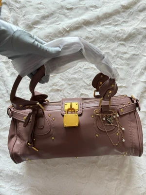 Rosa handväska med gulddetaljer - Säljer en snygg rosa handväska med guldfärgade metalldetaljer och dekorativa kedjor. Väskan har cylinderform, dubbla handtag och ett stort guldfärgat lås framtill. Perfekt accessoar för dig som gillar lyxig och unik design.