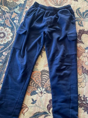 Mörkblå cargobyxor från Warp - Snygga mörkblå cargobyxor från Warp med elastisk midja och stora sidofickor. Byxorna är tillverkade i mjukt material och har en avslappnad passform som är perfekt för en chill look. Perfekt för dig som gillar streetstyle och funktionella plagg.